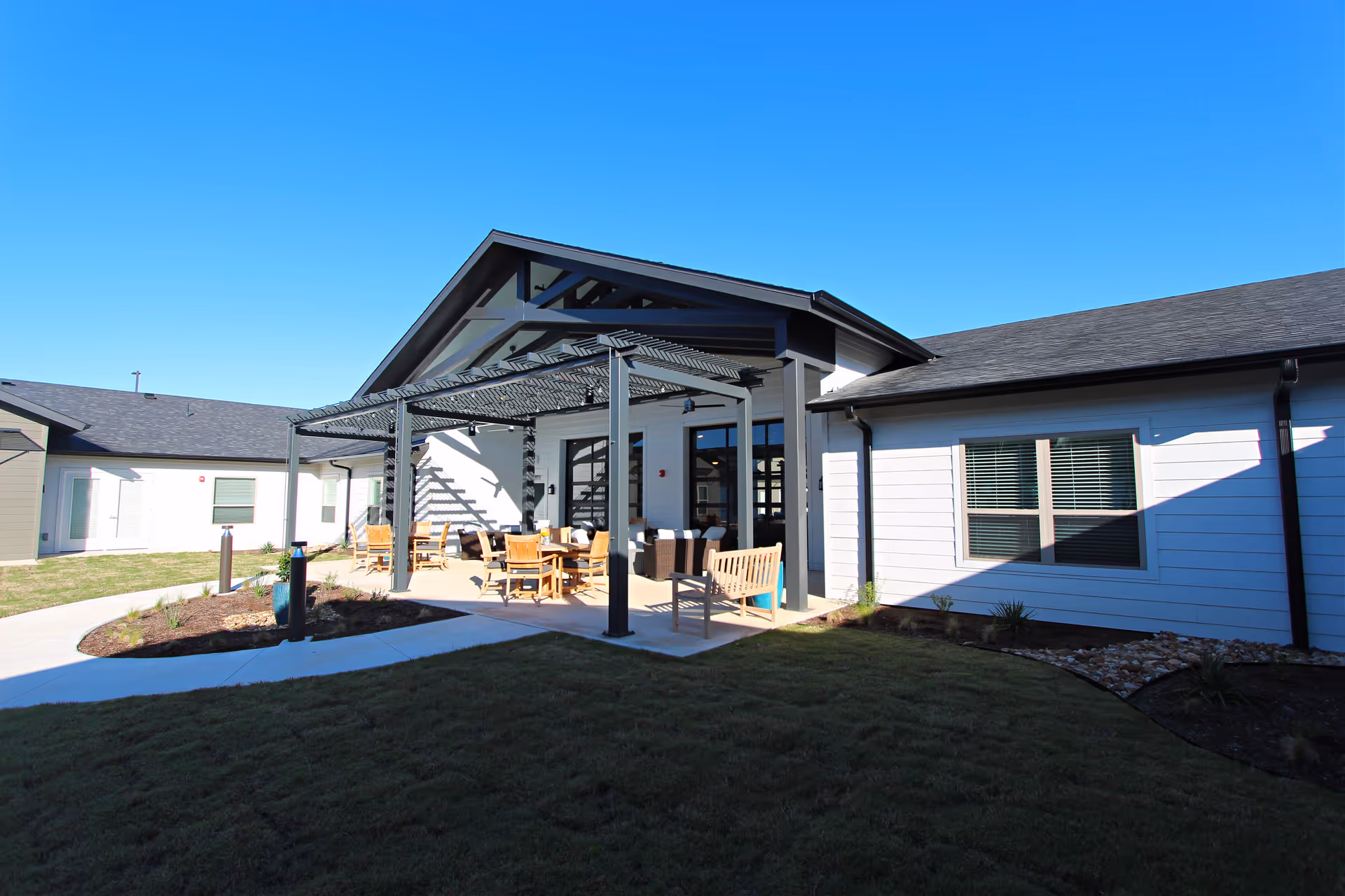 Exterior view of a single-story building with a covered patio, outdoor seating, and lawn under a clear blue sky.