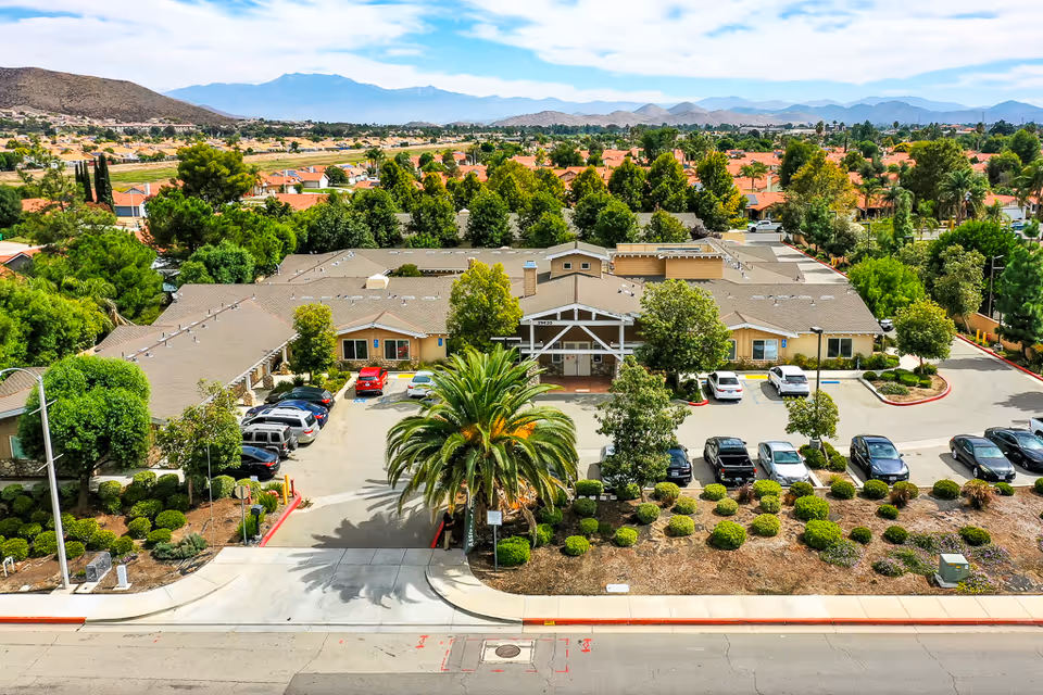Aerial view of a single-story senior living building with a central palm tree, parking lot, and surrounding homes and mountains.