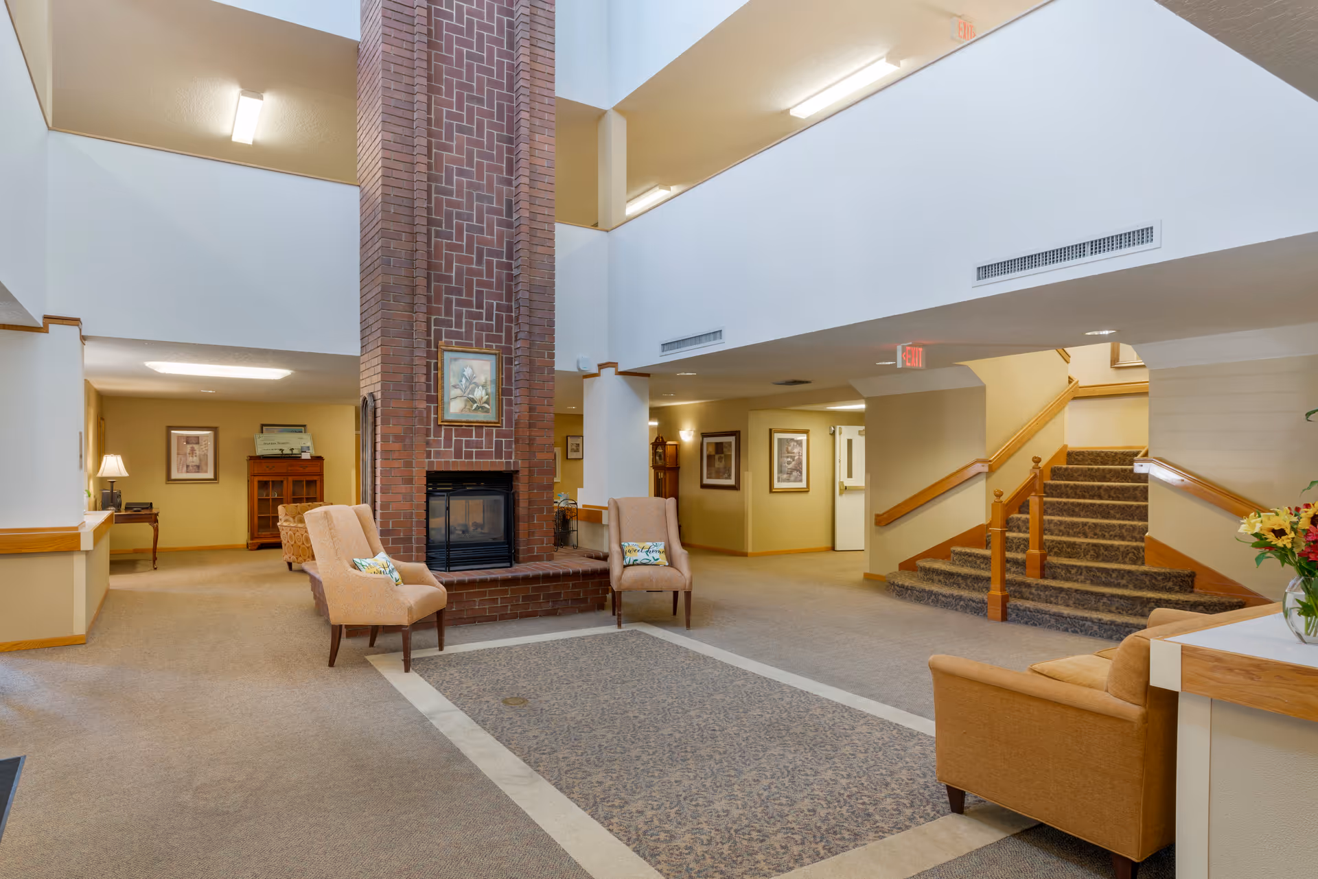 A spacious senior living facility common area with a tall brick fireplace in the center. The room has beige carpeting and walls, with several upholstered chairs arranged around the fireplace. There is a staircase with wooden handrails leading to an upper floor, framed artwork on the walls, and a vase with flowers on a table to the right.