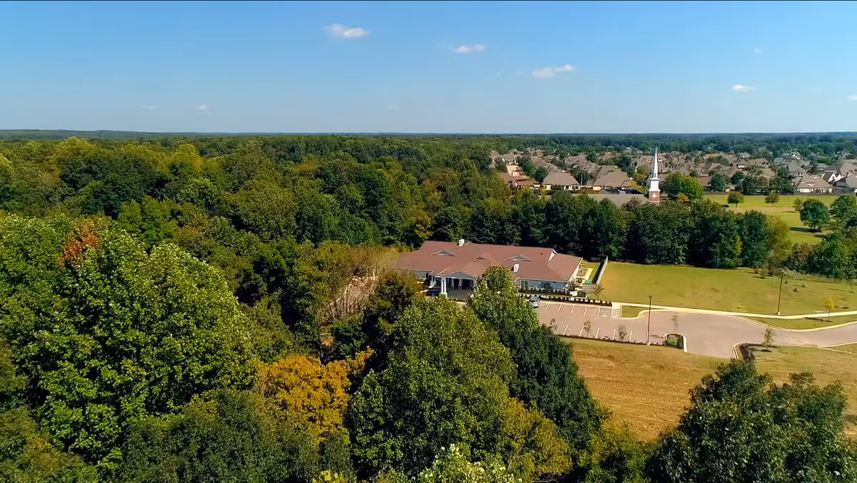 Aerial view of a senior living facility surrounded by dense green trees with a parking lot and a road nearby. In the background, there is a residential neighborhood and a church with a tall steeple under a clear blue sky.