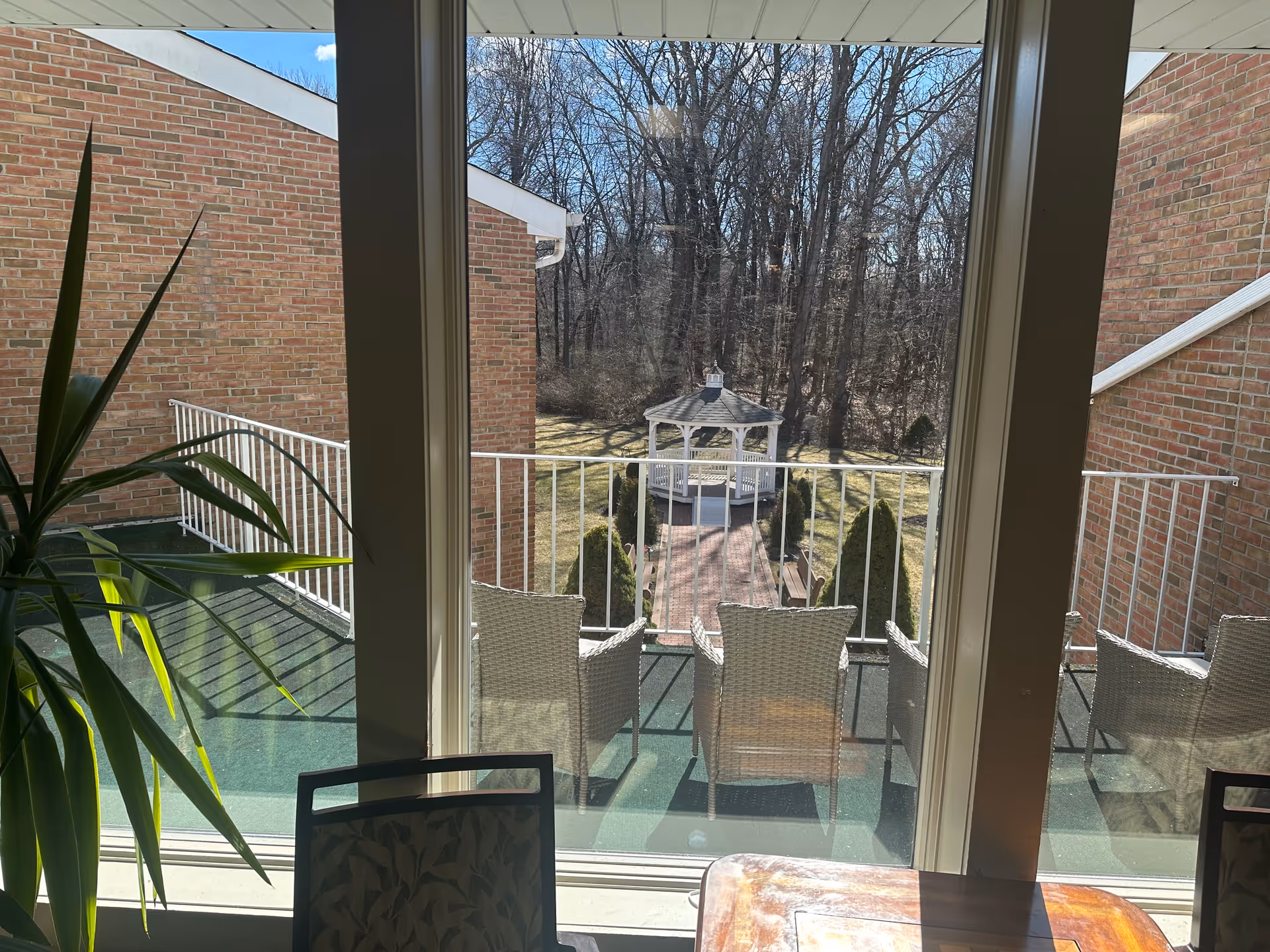 View through large glass doors showing a balcony with wicker chairs overlooking a garden area with a white gazebo and trees in the background. Part of a wooden table and a plant are visible inside near the doors.