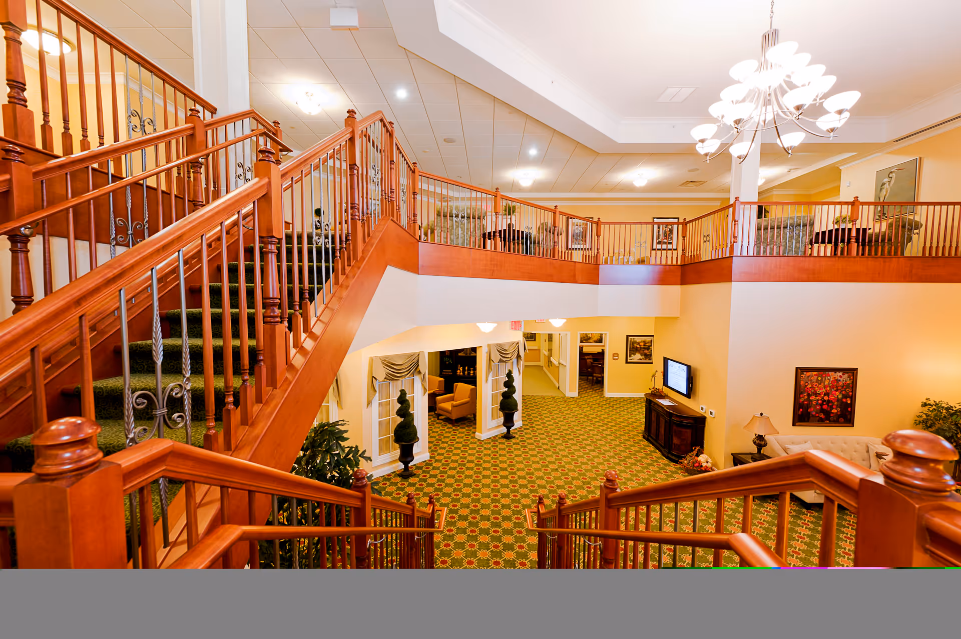 Interior view of a senior living facility featuring a large wooden staircase with green carpet leading to an upper balcony area. The space has warm yellow walls, patterned green carpet, a chandelier hanging from the ceiling, and various seating areas with chairs and a sofa. There are framed pictures on the walls and a TV mounted on a wooden cabinet.