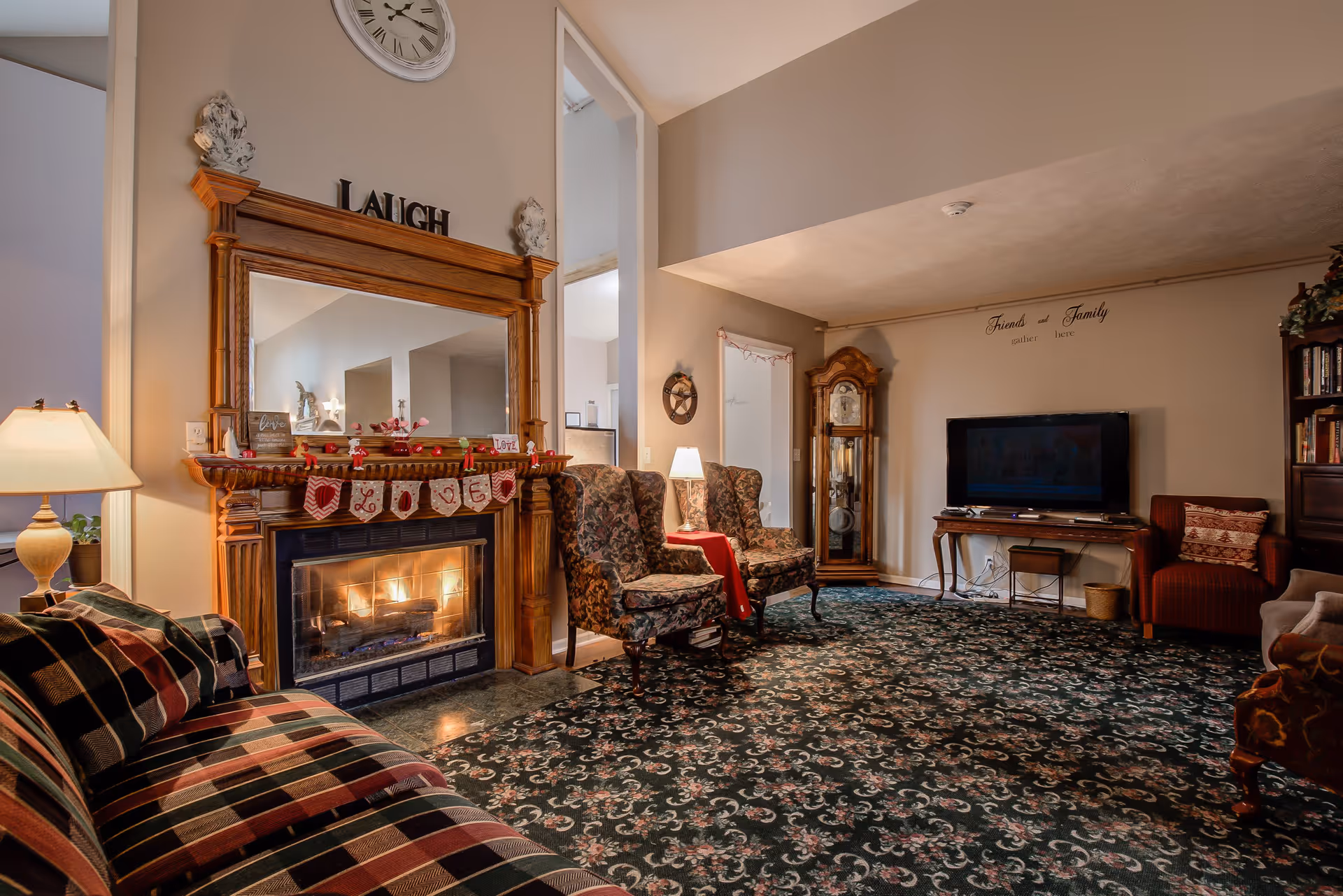 A cozy living room with a patterned carpet, a fireplace with a wooden mantel decorated with a LOVE banner, a large mirror above the fireplace with the word LAUGH on top, two floral upholstered armchairs next to a small table with a lamp, a grandfather clock, a flat-screen TV on a wooden table, and a bookshelf. The walls have decorative text that reads 'Friends and Family gather here.'