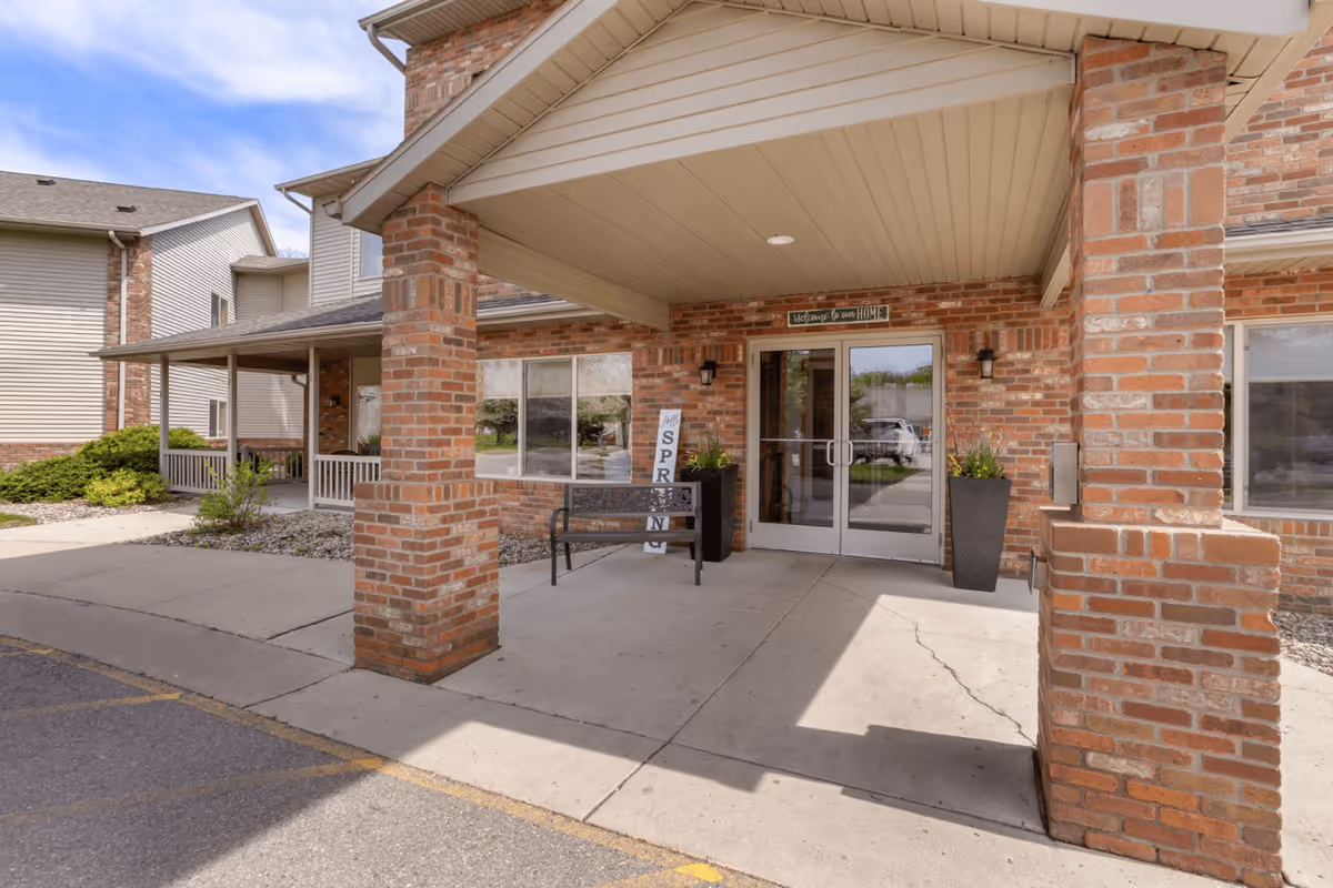 Entrance of a senior living facility with brick pillars and a covered porch. There is a bench, two tall planters with greenery, and a sign that says 'Hello SPRING'. Above the double glass doors, a sign reads 'Welcome to our HOME'.