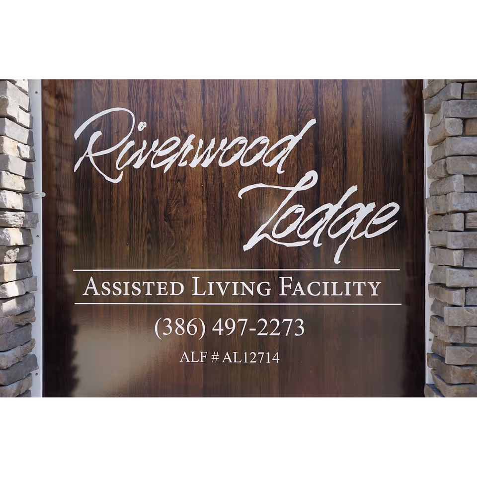 Wooden sign that reads "Riverwood Lodge Assisted Living Facility" with a phone number, flanked by stone columns.