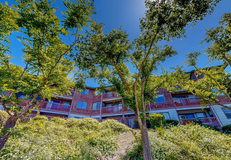 View of a multi-story residential building with red balconies partially obscured by green trees and bushes under a clear blue sky.
