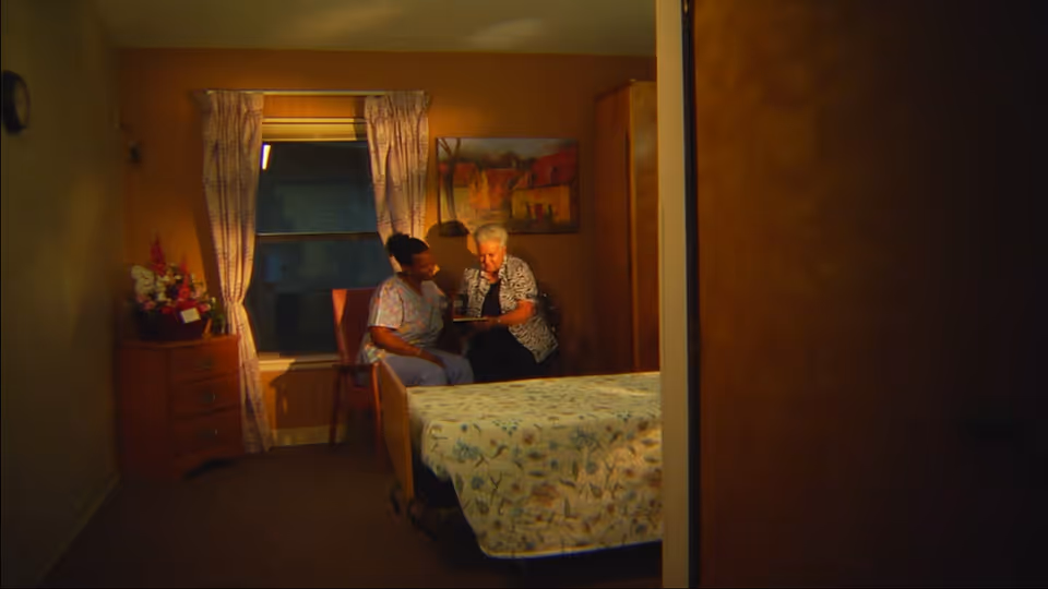 A cozy bedroom with a bed covered in a floral bedspread, a wooden dresser with flowers on top, and two women sitting on chairs near a window with patterned curtains. One woman appears to be a caregiver or nurse, and the other is an elderly woman. They are engaged in a conversation or activity together.
