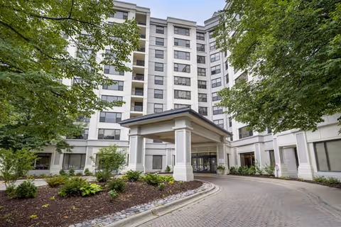 Exterior view of a multi-story senior living facility building with a covered entrance driveway, surrounded by landscaped greenery and trees.