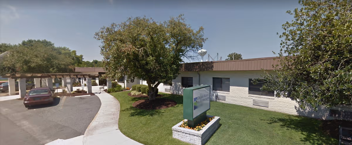 Exterior view of Delmar Nursing and Rehabilitation facility showing a single-story building with a brown roof, a green lawn with trees and shrubs, a sidewalk leading to the entrance, and a sign in front of the building.