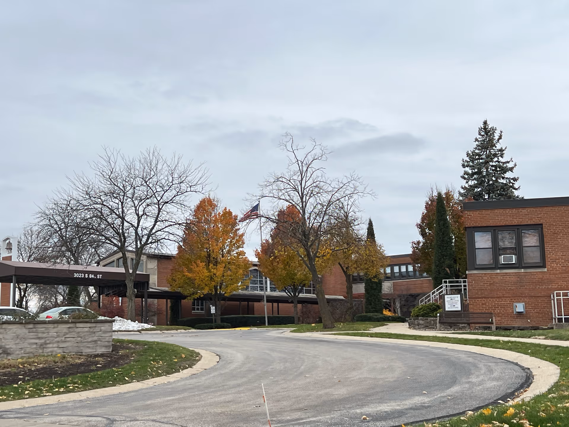 Exterior view of a brick building with a curved driveway in front. There are several trees with autumn foliage and some bare branches. An American flag is visible near the center of the image. The sky is overcast.