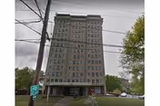Tall multi-story residential building with multiple windows, surrounded by trees and power lines in front. The sky is overcast.