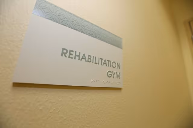 A close-up view of a wall sign that reads 'REHABILITATION GYM' with braille below the text, mounted on a beige textured wall.
