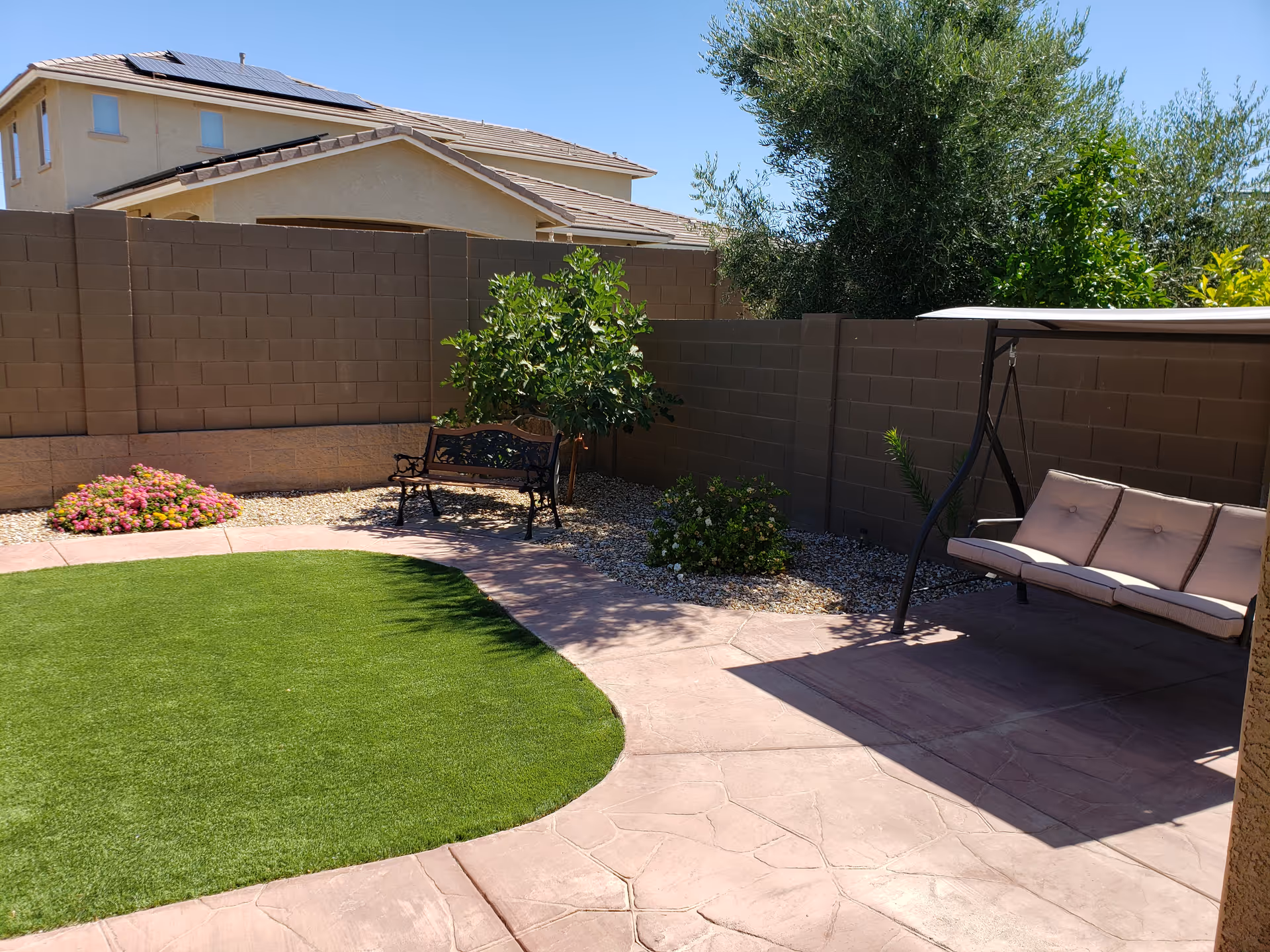 A sunny outdoor patio area with a curved patch of green artificial grass surrounded by a pinkish stone walkway. There is a black metal bench with decorative backrest near a small tree and some flowering plants along a brown brick wall. On the right side, there is a cushioned swing seat under a canopy. In the background, parts of neighboring houses and trees are visible under a clear blue sky.