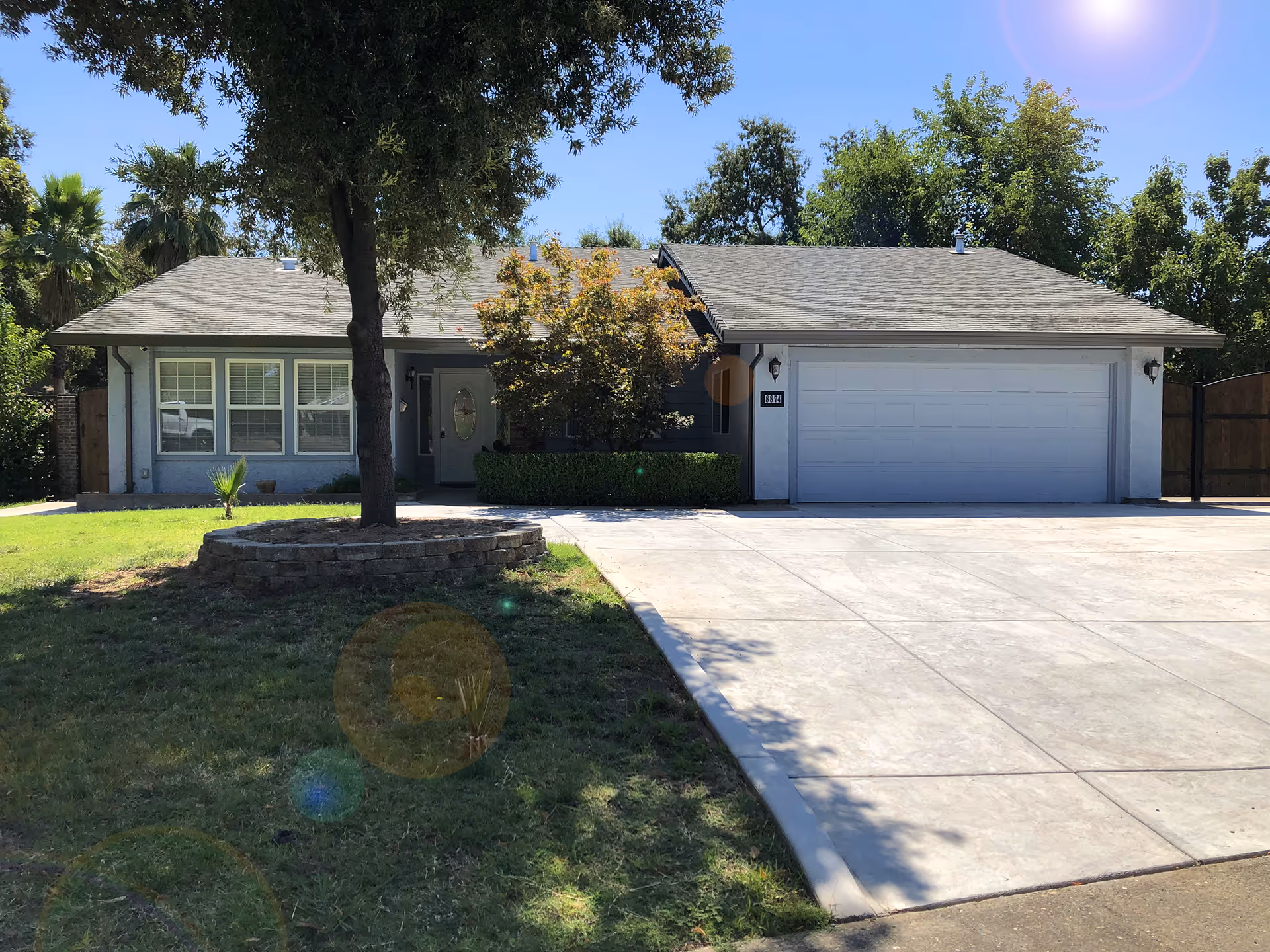 Front exterior view of a single-story house with a large driveway, a two-car garage, a tree surrounded by a circular brick planter in the front yard, and various trees and bushes around the property under a clear blue sky.