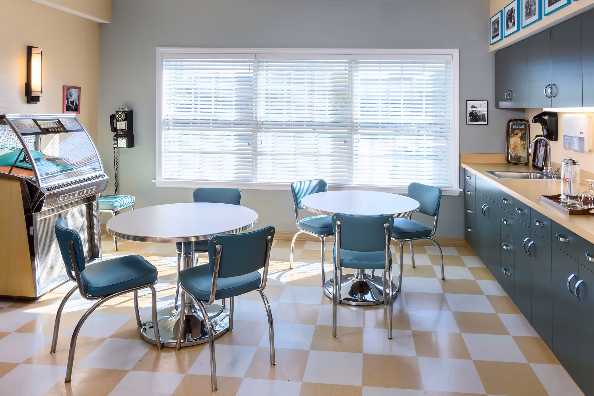 A bright retro-style dining area with two round white tables surrounded by teal chairs with chrome legs. The room features a checkered beige and white floor, a large window with white blinds, a vintage jukebox, a wall-mounted rotary phone, and a kitchenette with teal cabinets, a sink, and a coffee maker. Framed black and white photos decorate the walls.