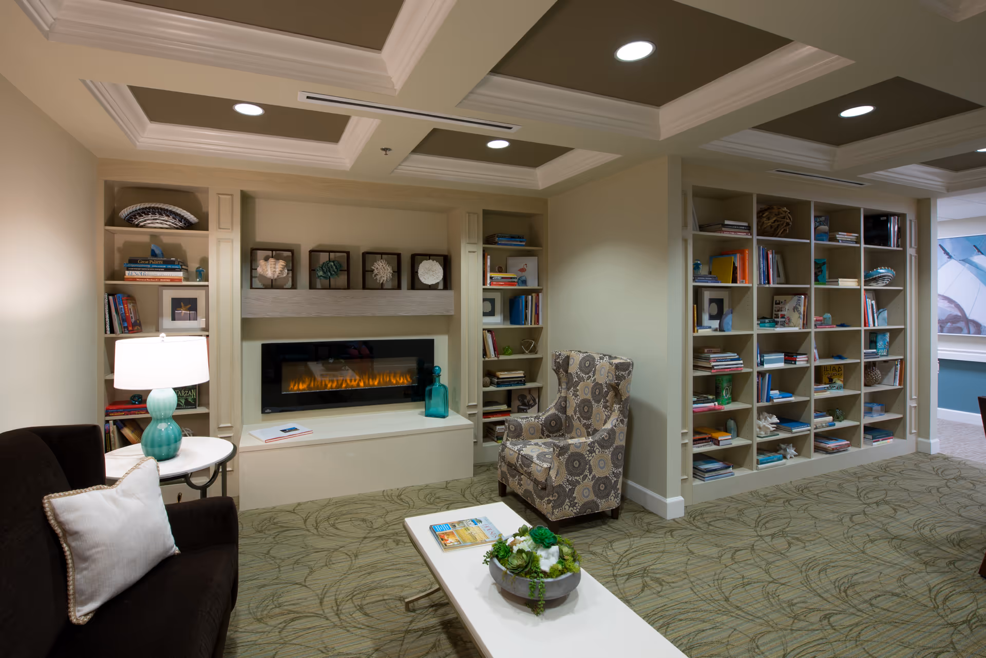 Cozy communal living room with built-in bookshelves, a modern fireplace, patterned armchair, sofa, and coffee table.