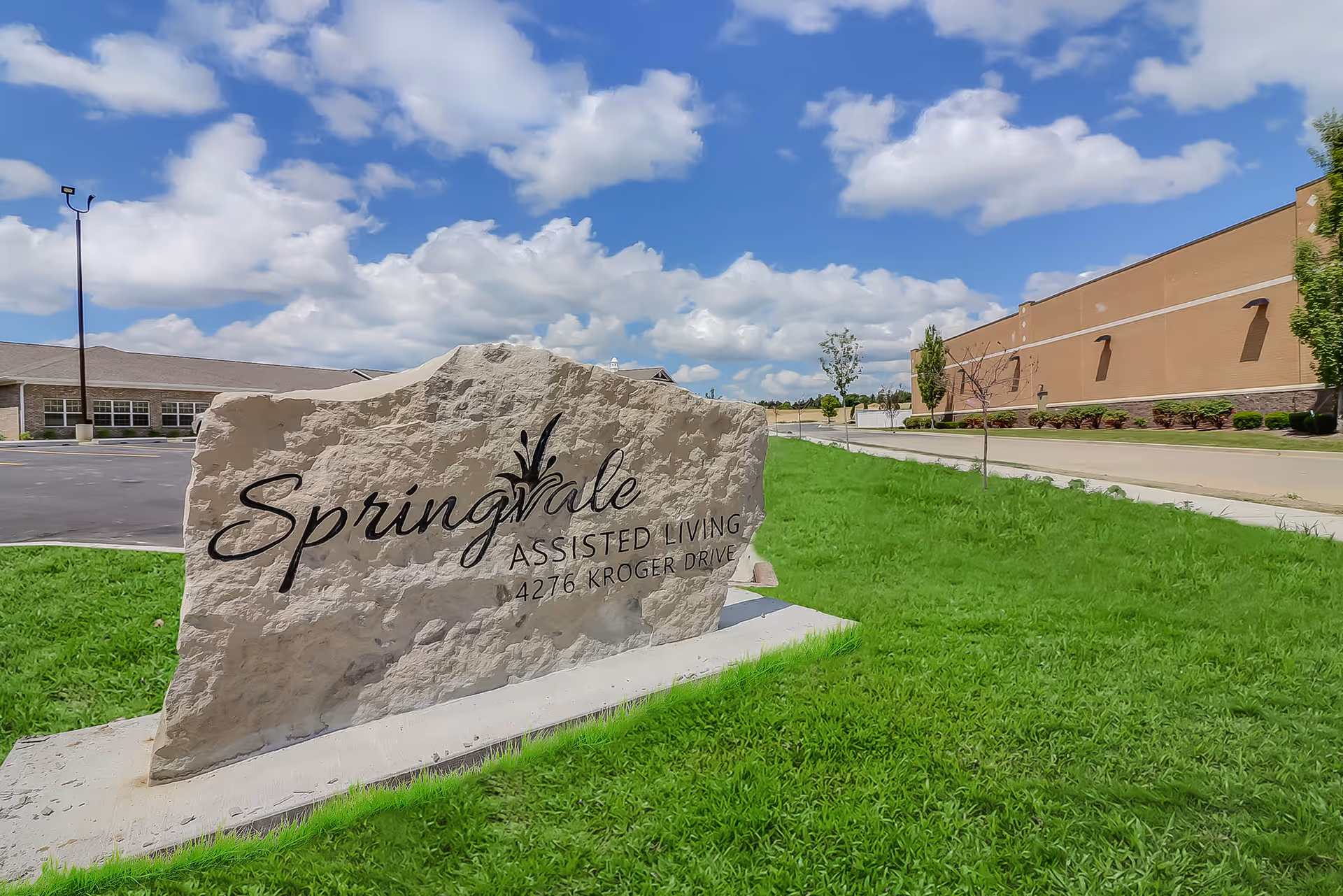 Stone entrance sign reading 'Springvale Assisted Living' on a grassy lawn with the facility buildings and a blue sky in the background.