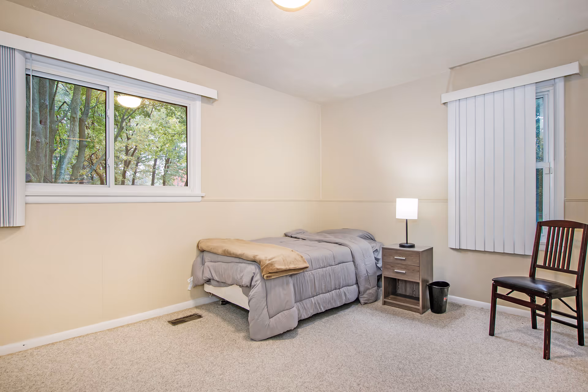 A simple bedroom with beige walls and carpeted floor. There is a single bed with gray bedding and a tan blanket folded at the foot. Next to the bed is a small wooden nightstand with a lit lamp and a black trash bin underneath. A wooden chair with a dark finish is placed near a window with vertical blinds. Another window with vertical blinds shows green trees outside.