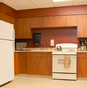 A kitchen area with wooden cabinets, a white refrigerator on the left, a white stove with a striped towel hanging on the handle, a microwave on the countertop, a paper towel dispenser, and a soap dispenser mounted on the wall above the sink.