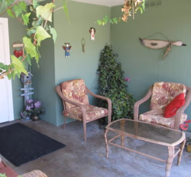 A cozy outdoor seating area with two cushioned wicker chairs featuring floral patterns, a glass-top wicker coffee table, green walls decorated with hanging ornaments, and some potted plants including a leafy vine hanging from above.