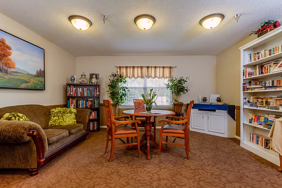 A cozy common area in a senior living facility featuring a round wooden table with four chairs, two large potted plants near a window with a valance, a brown upholstered sofa with green pillows, bookshelves filled with books, a painting of a landscape on the wall, and a cabinet with a blue cloth and decorative items.