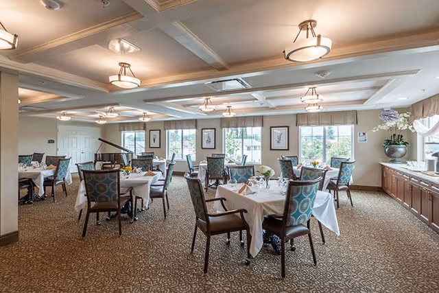 A spacious dining room with multiple tables covered in white tablecloths, each set with glassware and floral centerpieces. The room features patterned carpet, large windows letting in natural light, framed artwork on the walls, and ceiling light fixtures. There is a piano in the corner and a long counter with cabinets and decorative plants along one side.