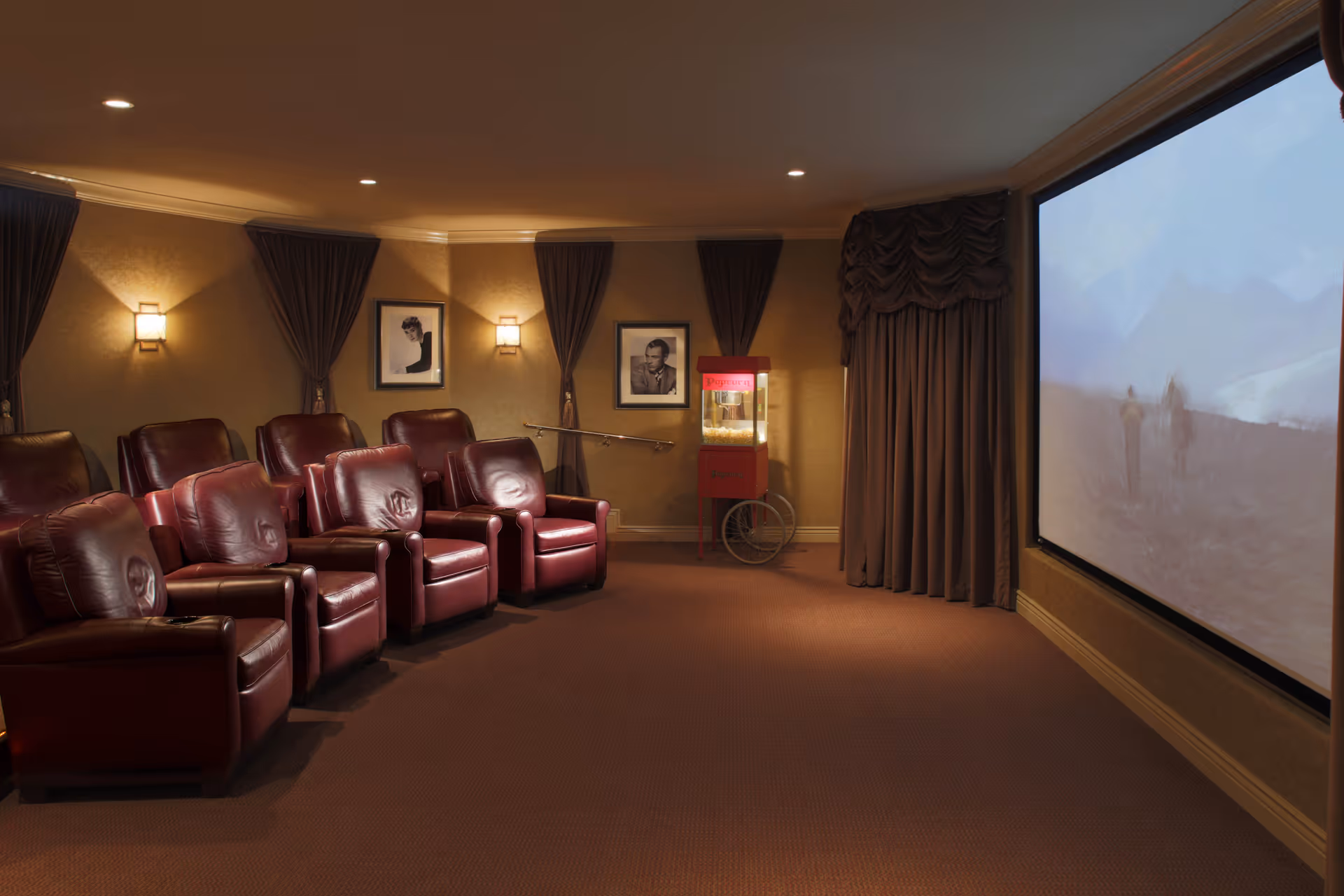 A cozy home theater room with two rows of red leather recliner chairs facing a large projection screen. The walls are decorated with framed black and white portraits and wall sconces emitting warm light. A vintage-style popcorn machine is positioned near the screen, and heavy curtains frame the screen area.