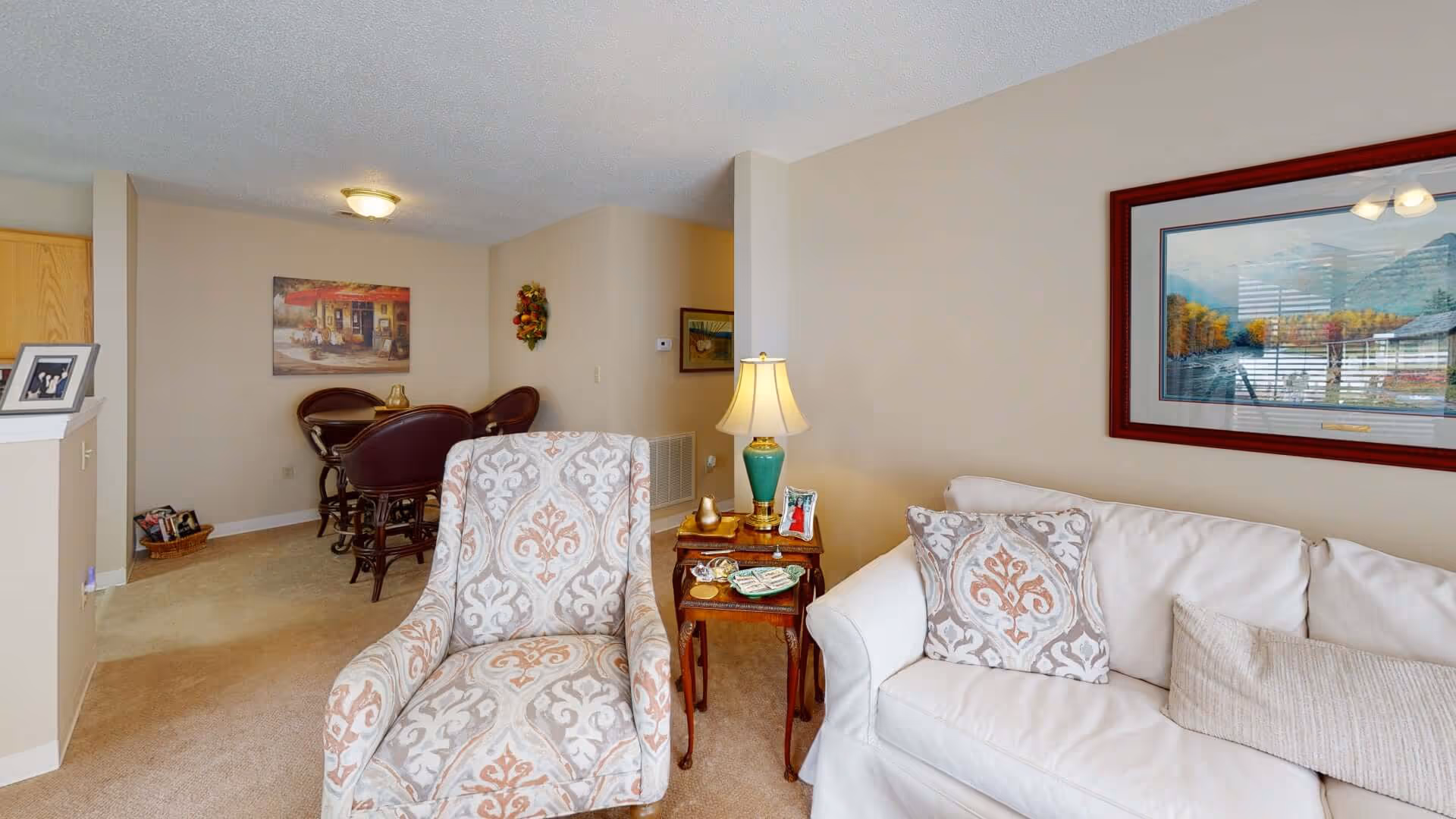 A cozy living room area with a patterned armchair and a white couch with decorative pillows. A small wooden side table with a green lamp, framed photo, and decorative items sits between the chair and couch. In the background, there is a dining area with a round table and four chairs, a wall painting, and beige walls and carpet.