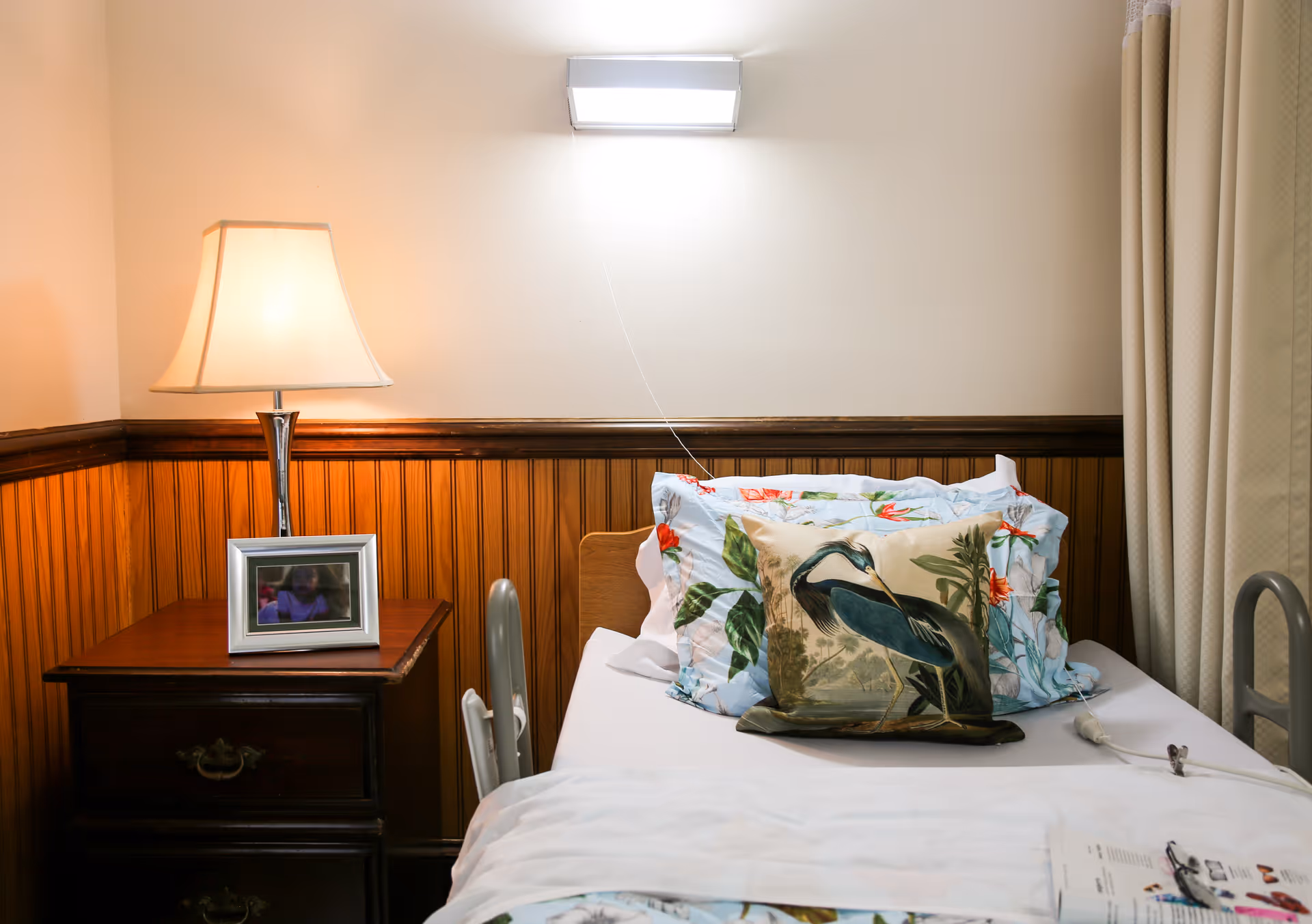 A cozy bedroom corner in a senior living facility featuring a bed with floral and bird-themed pillows, a wooden nightstand with a table lamp and a framed photo, and beige curtains on the right side.