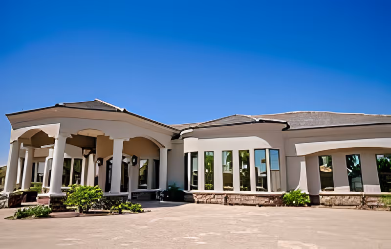 Exterior view of a single-story building with large windows and white columns under a clear blue sky, surrounded by some greenery and a paved area in front.