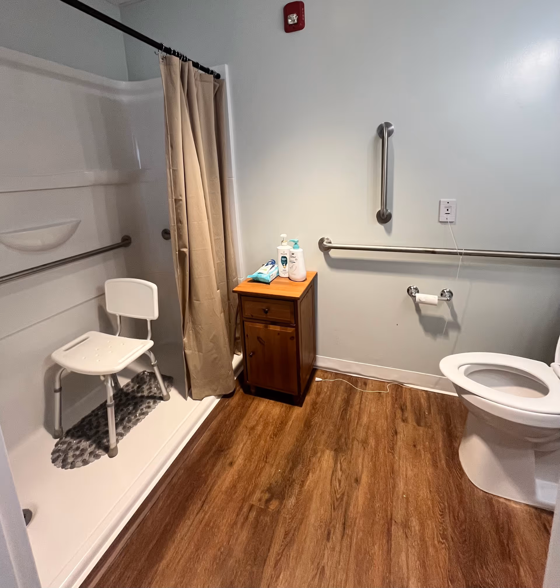 A bathroom with a walk-in shower featuring a white shower chair and beige shower curtain. There are grab bars on the walls near the shower and toilet for accessibility. A small wooden cabinet holds toiletries including lotion and wipes. The floor has wood-like vinyl flooring.