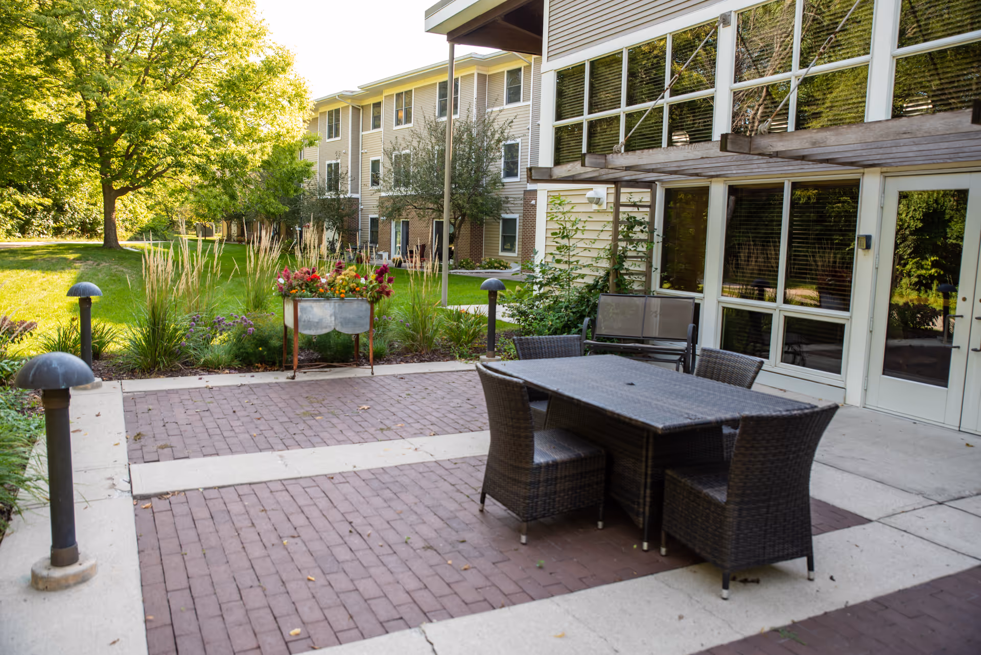 A patio with a wicker dining table and chairs, planter with flowers, and grassy courtyard outside a senior living building.
