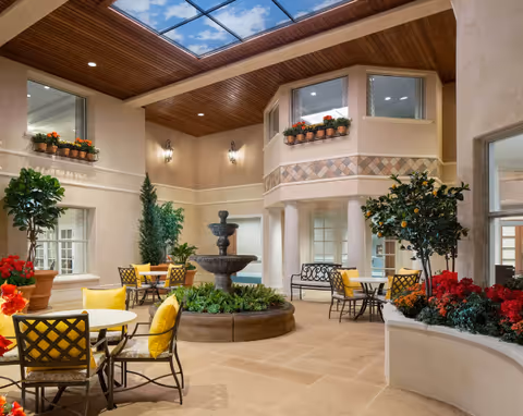 Indoor courtyard area with a central water fountain surrounded by greenery and potted plants. Several round tables with yellow cushioned chairs are arranged around the space. The ceiling features a large skylight with a wooden finish, allowing natural light to illuminate the area. The walls have windows with flower boxes and decorative wall sconces.