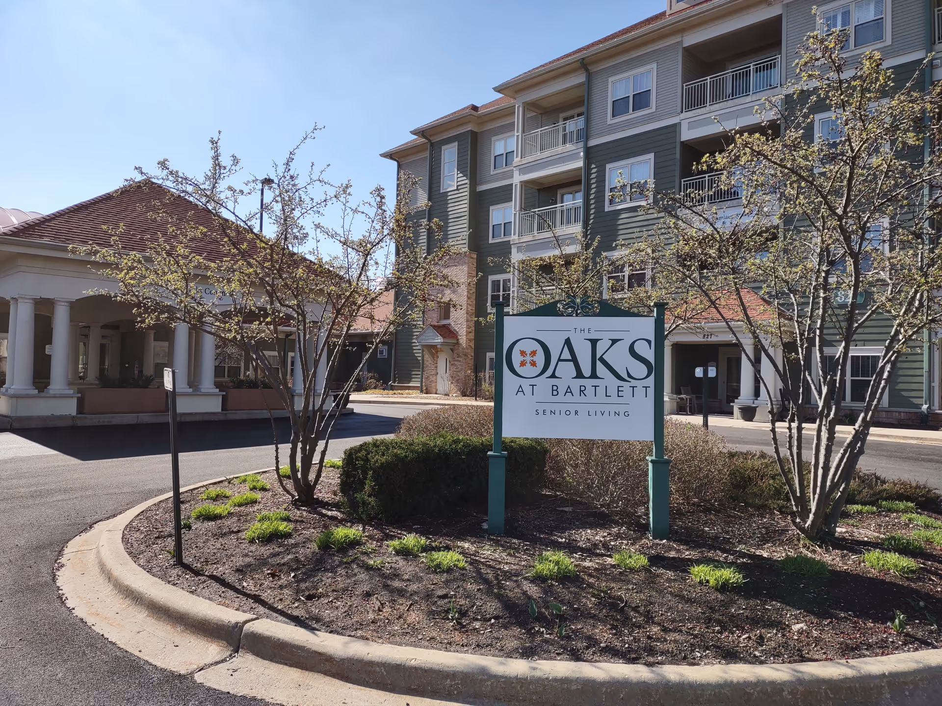 Exterior view of The Oaks at Bartlett senior living facility showing a multi-story building with balconies, a covered entrance with columns, and a landscaped area with trees and bushes surrounding a sign that reads 'The Oaks at Bartlett Senior Living'.