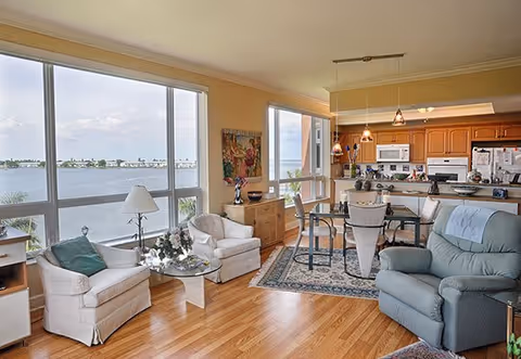 A bright and cozy living area with large windows offering a scenic view of a body of water. The room features two white armchairs with a glass coffee table between them, a blue recliner, a dining table with chairs on a patterned rug, and a kitchen area with wooden cabinets and white appliances in the background.