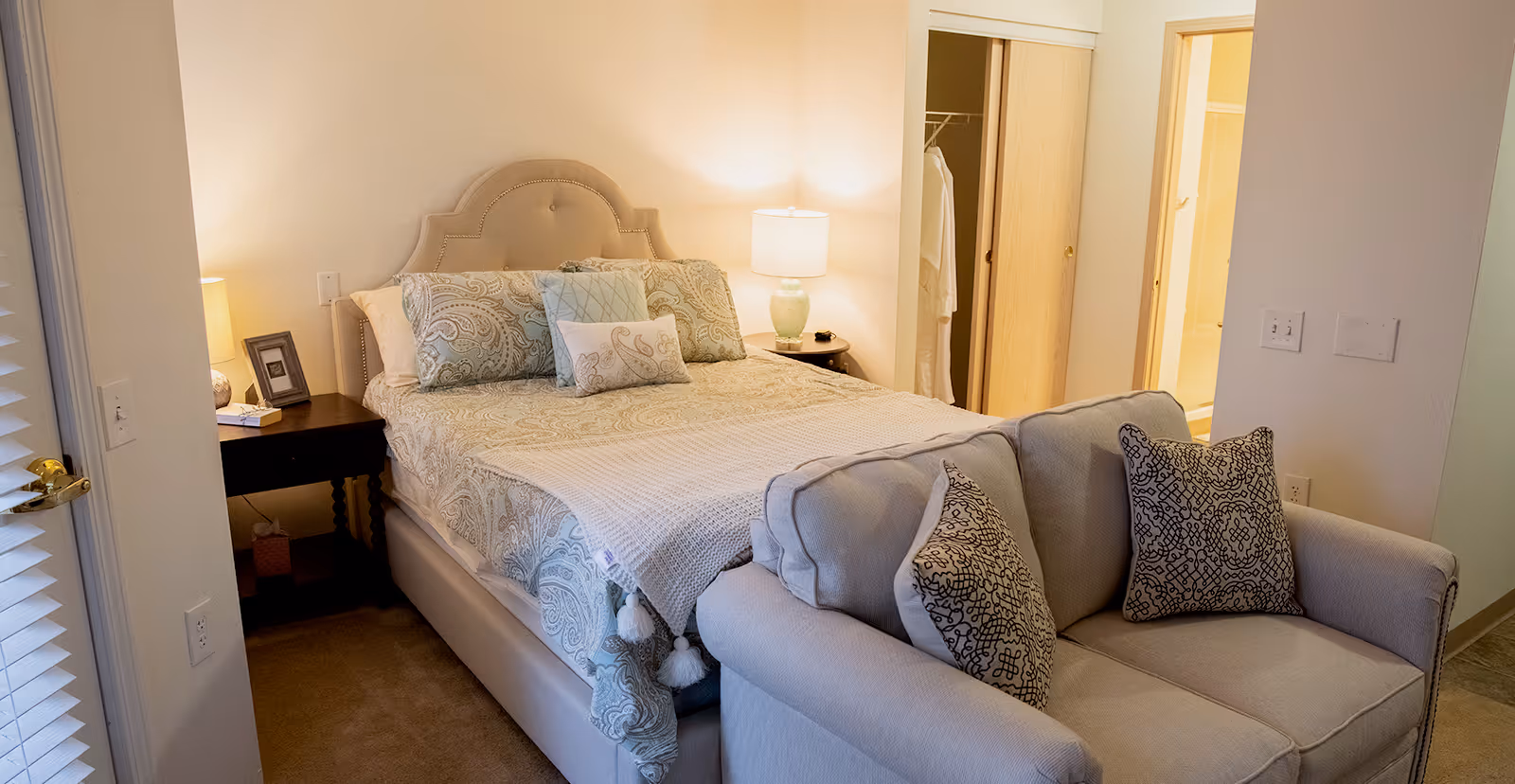 A cozy bedroom with a neatly made bed featuring patterned bedding and multiple pillows. There is a small sofa with decorative pillows at the foot of the bed. Two bedside tables with lamps flank the bed. A closet with sliding doors and an open bathroom door are visible in the background.