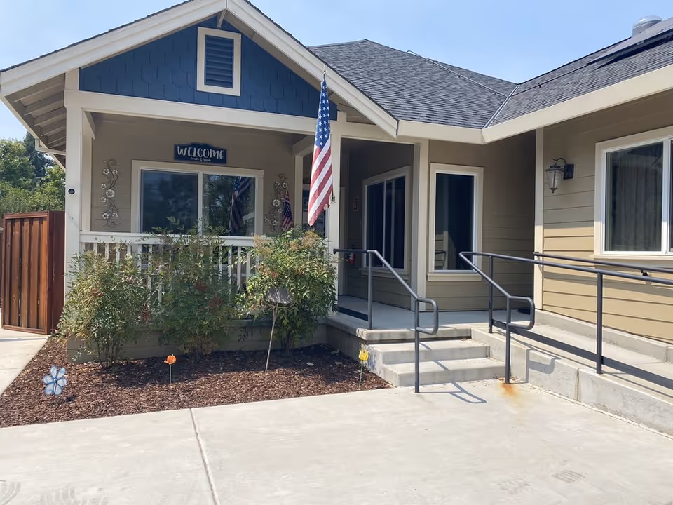 Exterior view of a single-story building with beige siding and a dark blue gable. There is a small porch with a white railing, bushes in front, and an American flag hanging near the entrance. A welcome sign is mounted above the window on the porch. The entrance has steps and a ramp with handrails leading up to it.