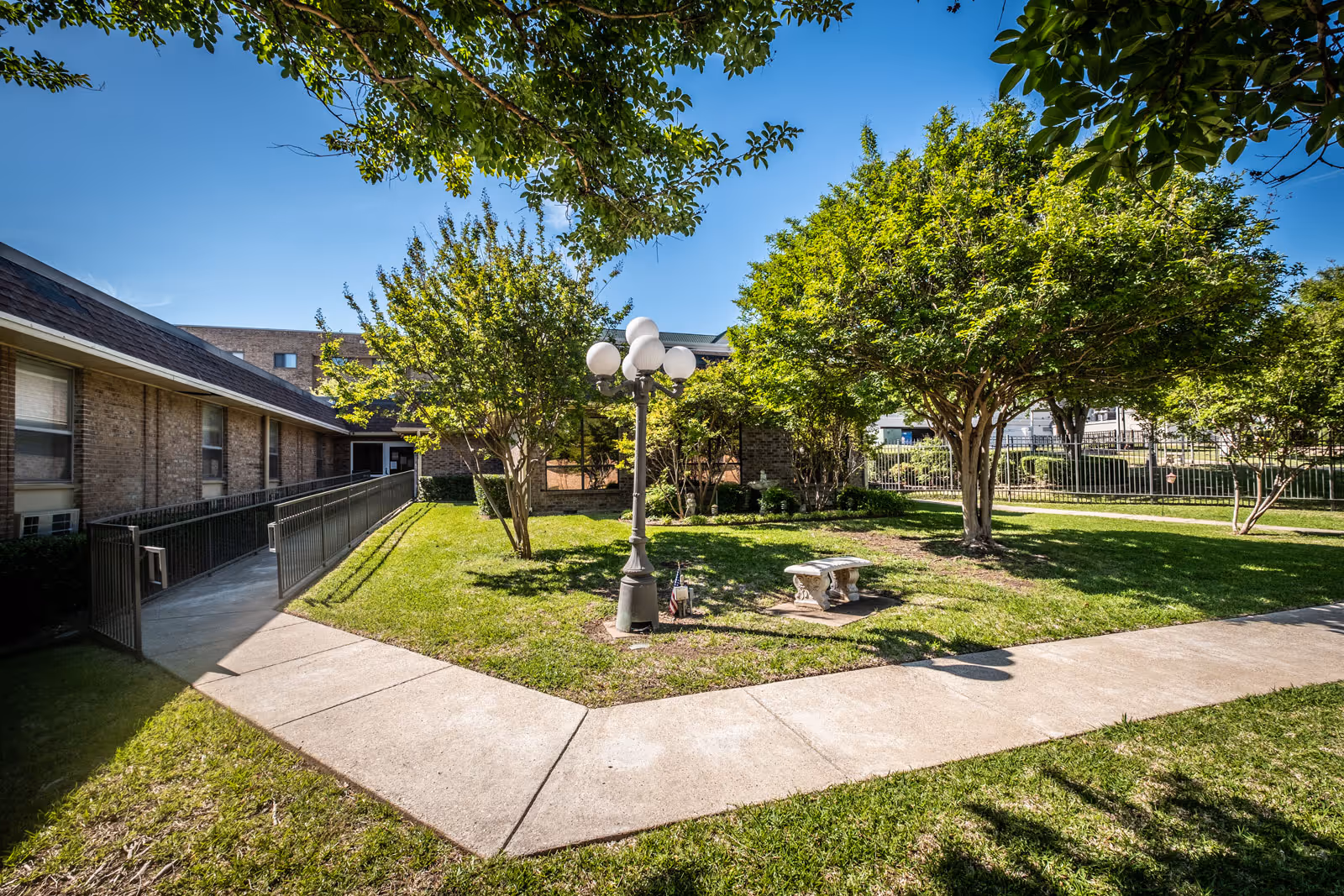 Outdoor courtyard area at Christian Care Nursing & Rehabilitation featuring a concrete pathway, green grass, several trees, a decorative street lamp with multiple globes, and a stone bench. The courtyard is surrounded by a brick building and a metal fence under a clear blue sky.