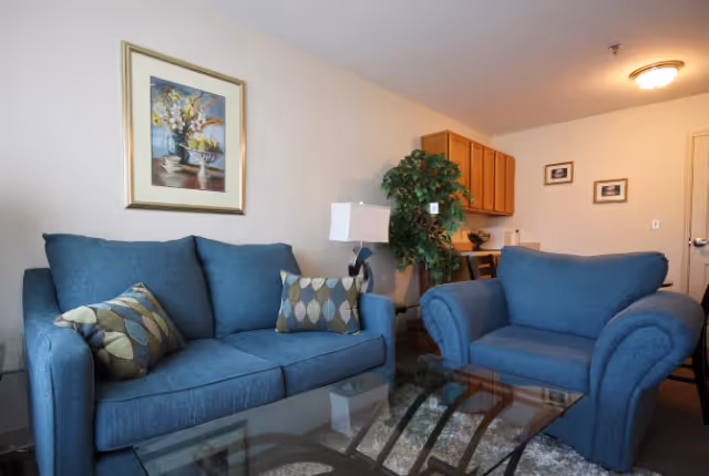 Cozy living room with a blue sofa and armchair, glass coffee table, framed artwork, and a small kitchenette visible in the background.