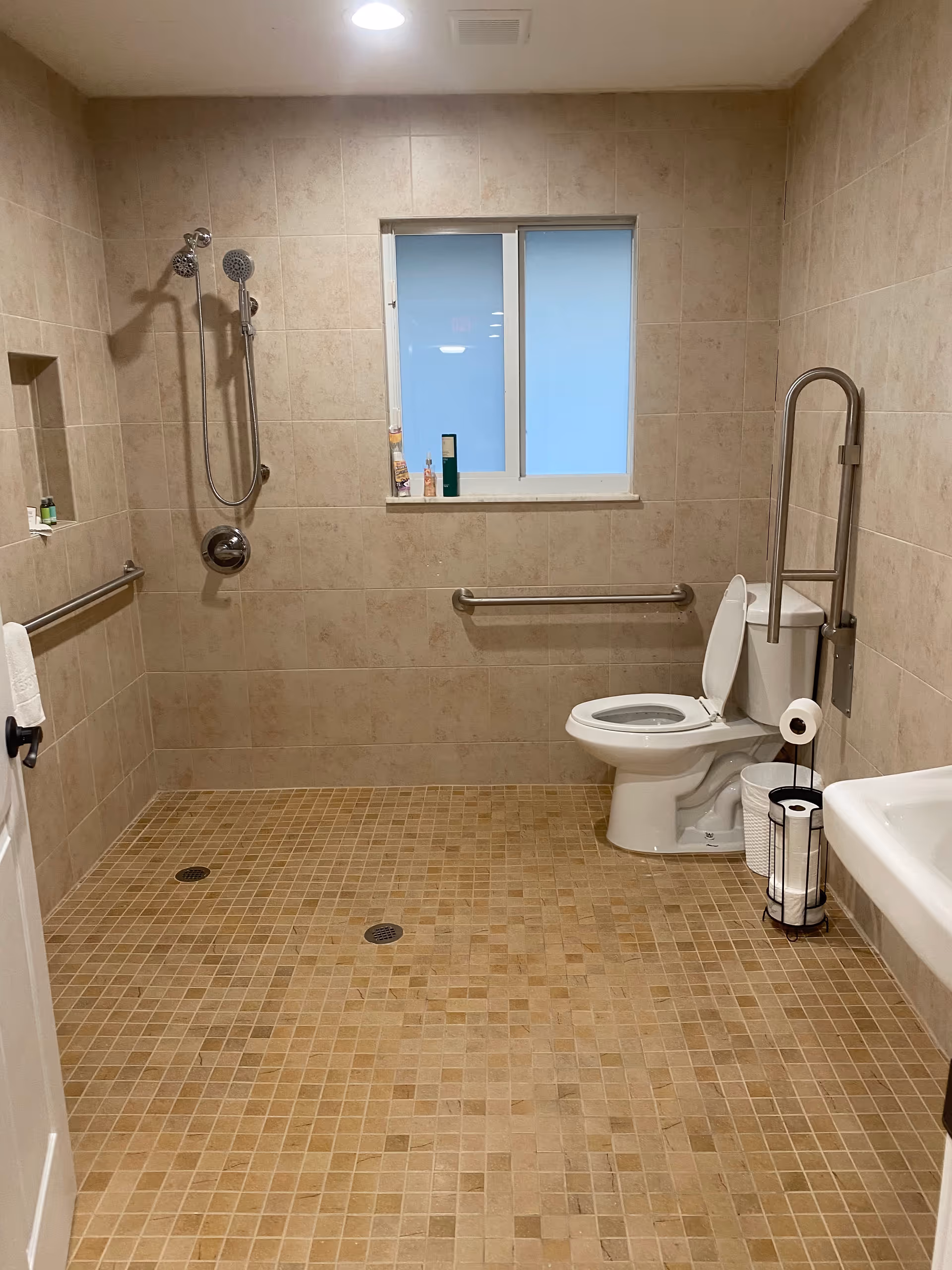 Accessible tiled bathroom with a roll-in shower, grab bars, toilet with a support rail, and a wall-mounted sink.