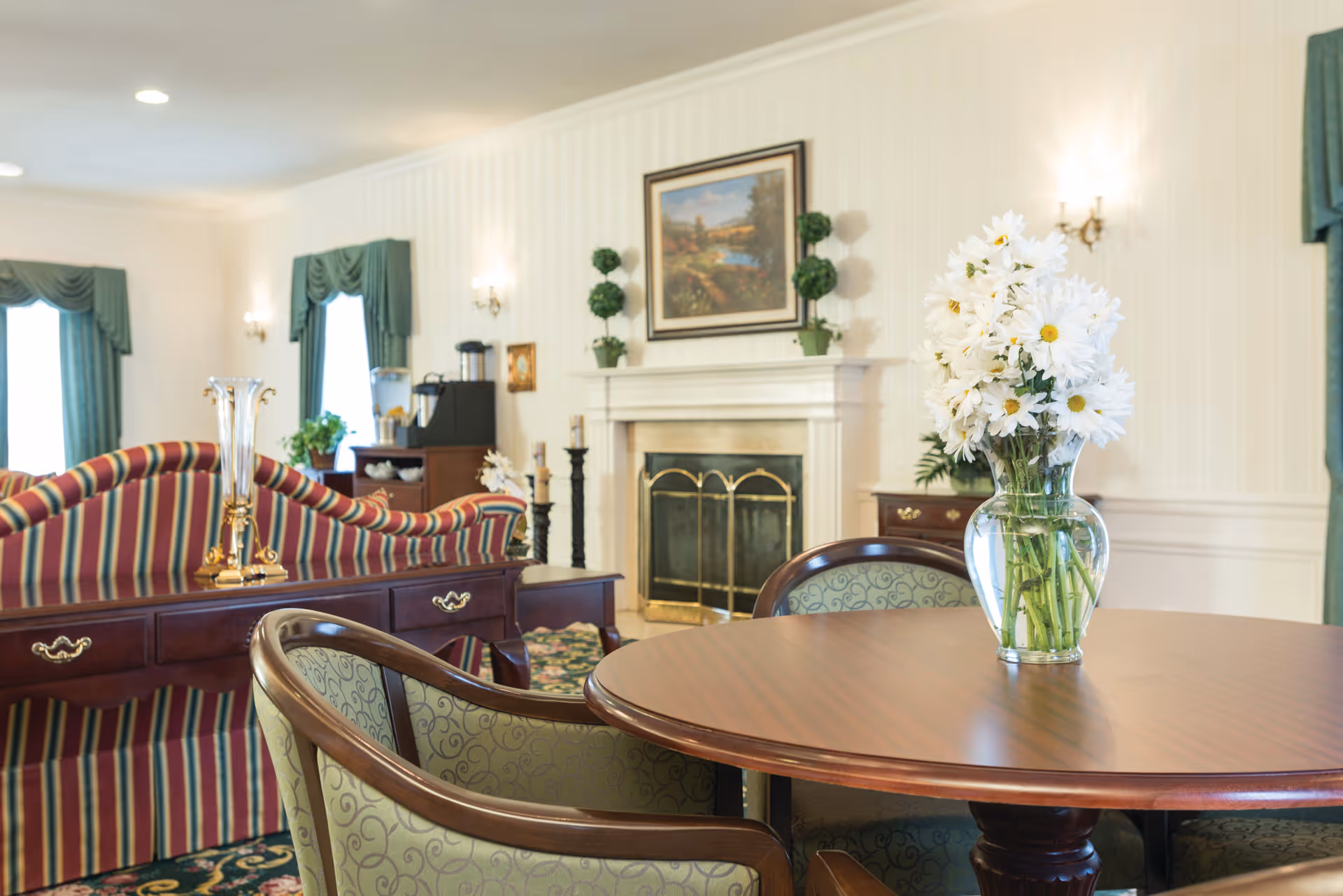 A cozy living room area with a round wooden table in the foreground holding a glass vase filled with white daisies. Behind the table are upholstered chairs with green patterned fabric. In the background, there is a striped sofa with red, green, and yellow colors, a wooden sideboard with decorative items, and a fireplace with a brass screen. The walls are light-colored with elegant sconces and green curtains framing the windows.