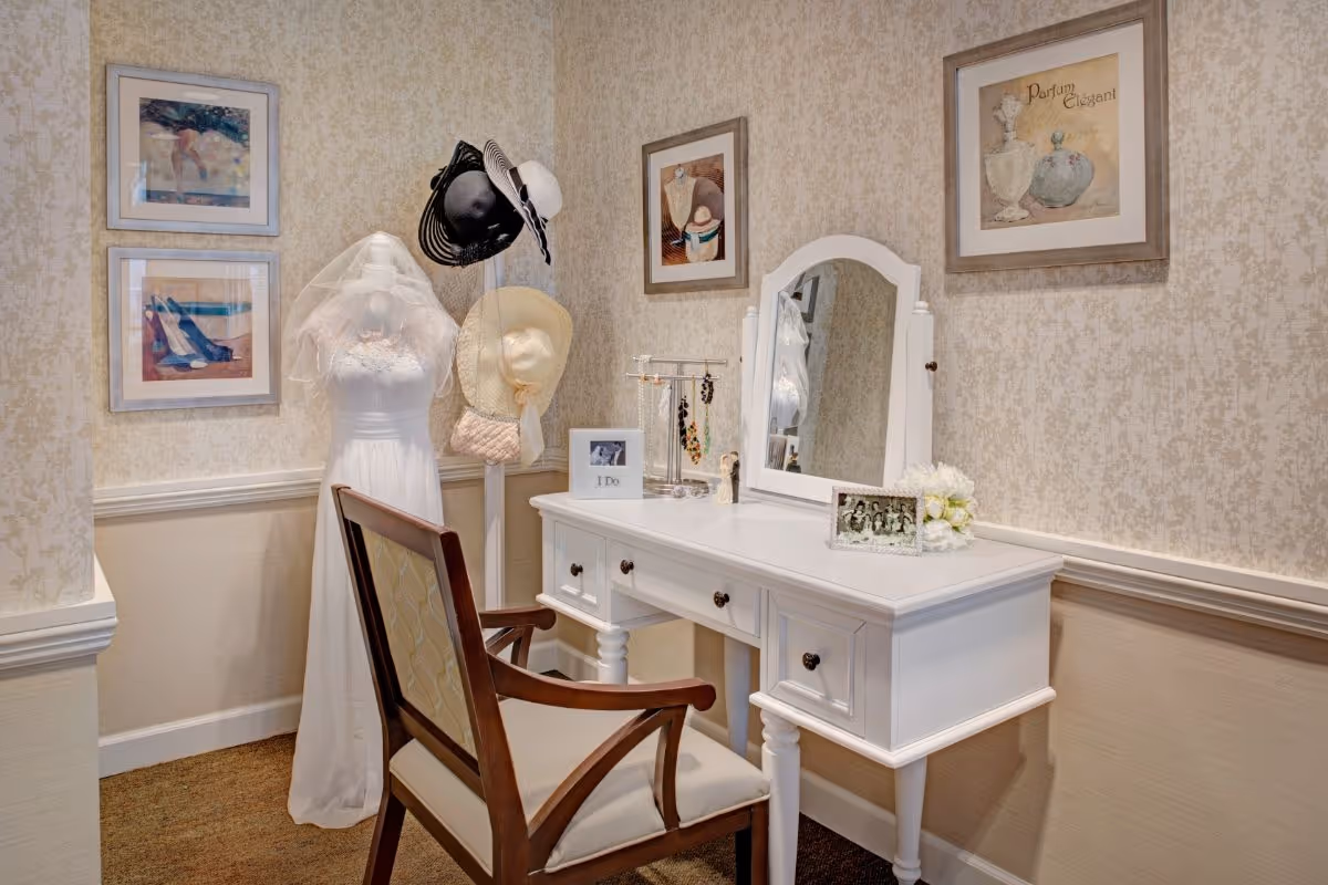 A white vanity table with mirror and chair beside a mannequin wearing a wedding dress and hats in a decorated dressing nook.