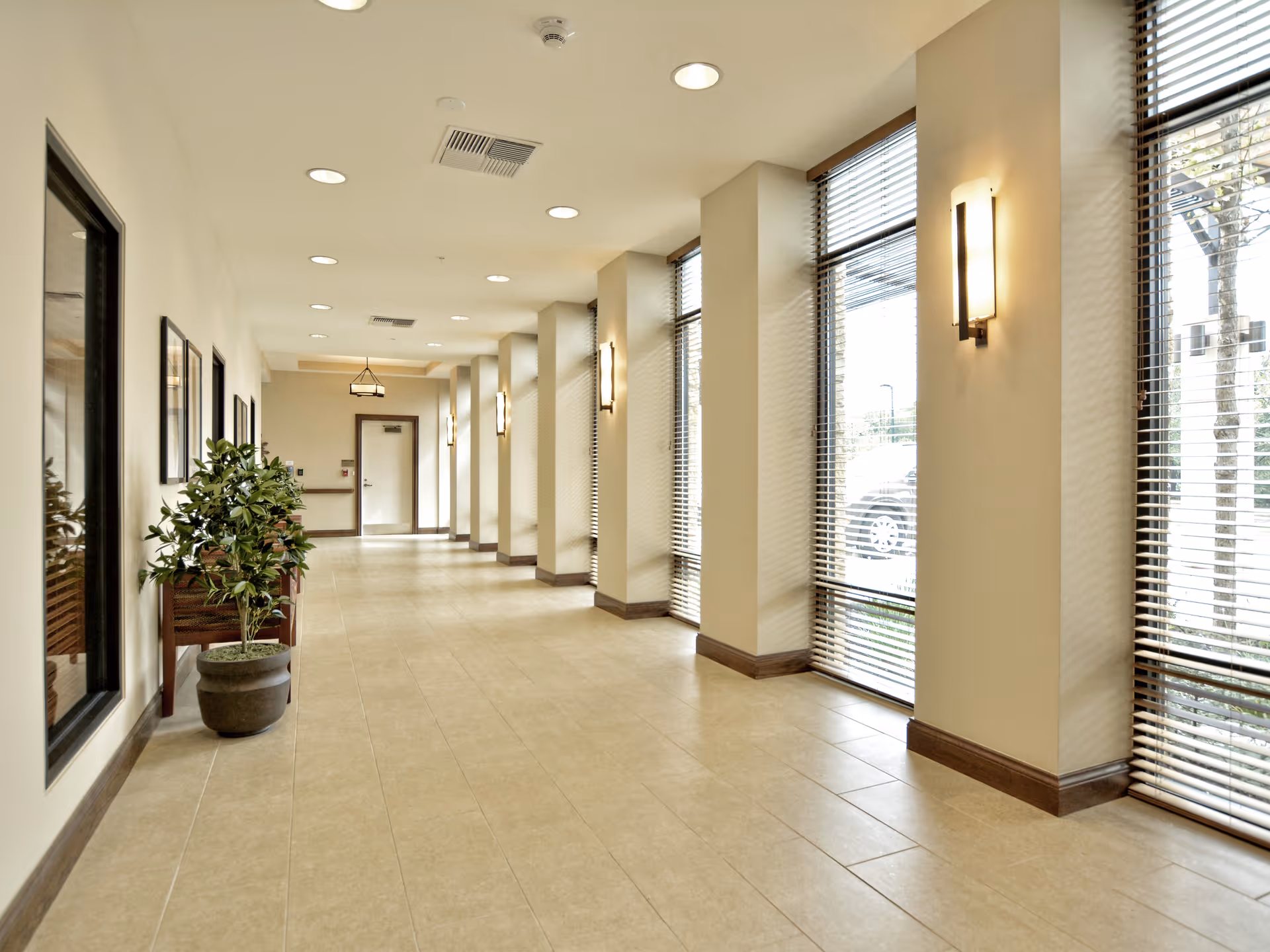 Bright, spacious hallway with tall windows, blinds, wall sconces, and a potted plant.