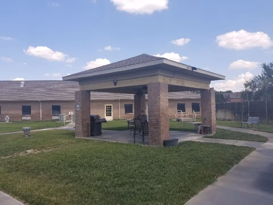 Covered brick pavilion with chairs and a grill in a grassy courtyard of a nursing facility under a blue sky.
