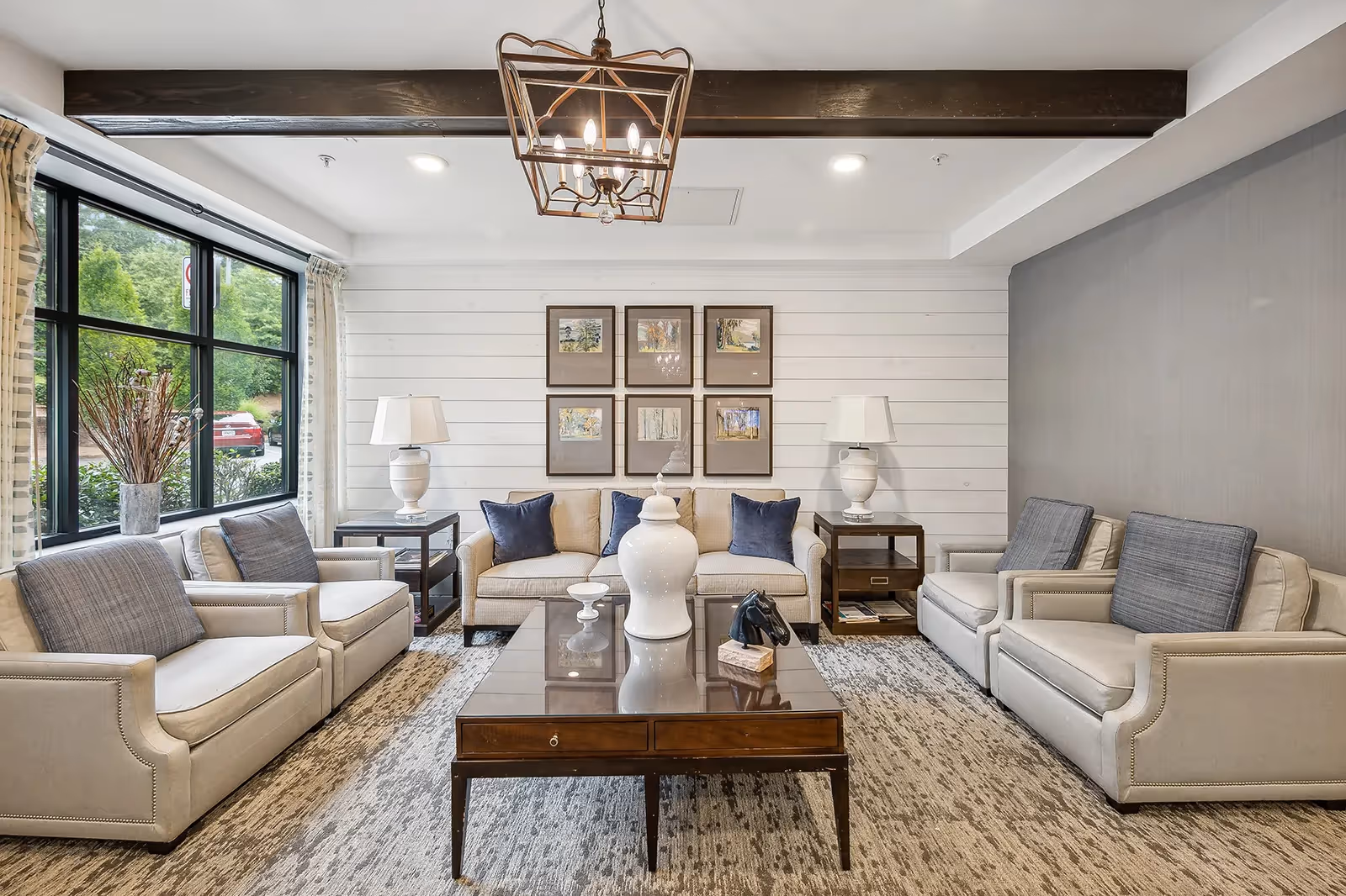 Bright modern living room with beige sofas and armchairs arranged around a dark wood coffee table beneath a decorative chandelier and large window.