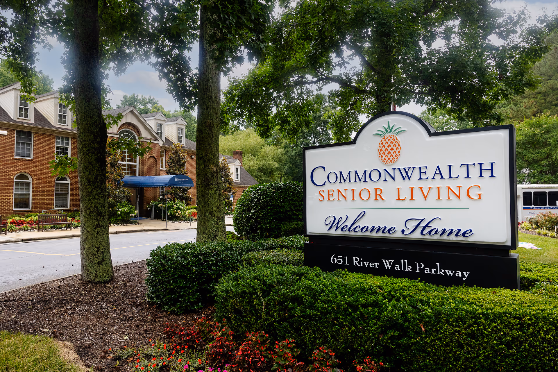Entrance sign for Commonwealth Senior Living at Georgian Manor with a large white sign featuring a pineapple logo and the text 'Commonwealth Senior Living Welcome Home 651 River Walk Parkway' surrounded by green bushes and trees, with a brick building and a blue awning in the background.