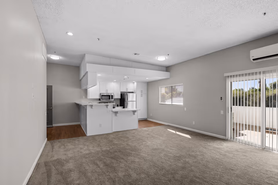 An empty open-concept living space in a senior living community featuring a carpeted living area, a kitchen with white cabinetry, stainless steel appliances including a refrigerator and microwave, a window with blinds, and a sliding glass door leading to an outdoor patio. The walls are painted light gray and the ceiling has recessed lighting and a mounted air conditioning unit.