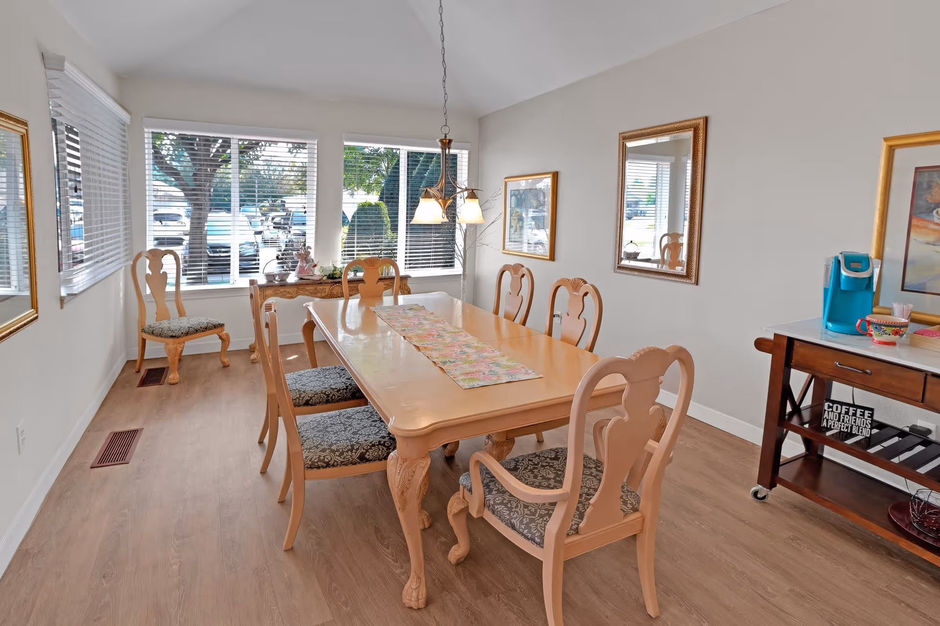 A bright dining room with a wooden dining table surrounded by six chairs with patterned cushions. Large windows with white blinds let in natural light, and there are framed pictures and a mirror on the walls. A wooden side table with a coffee maker and cups is on the right side of the room.