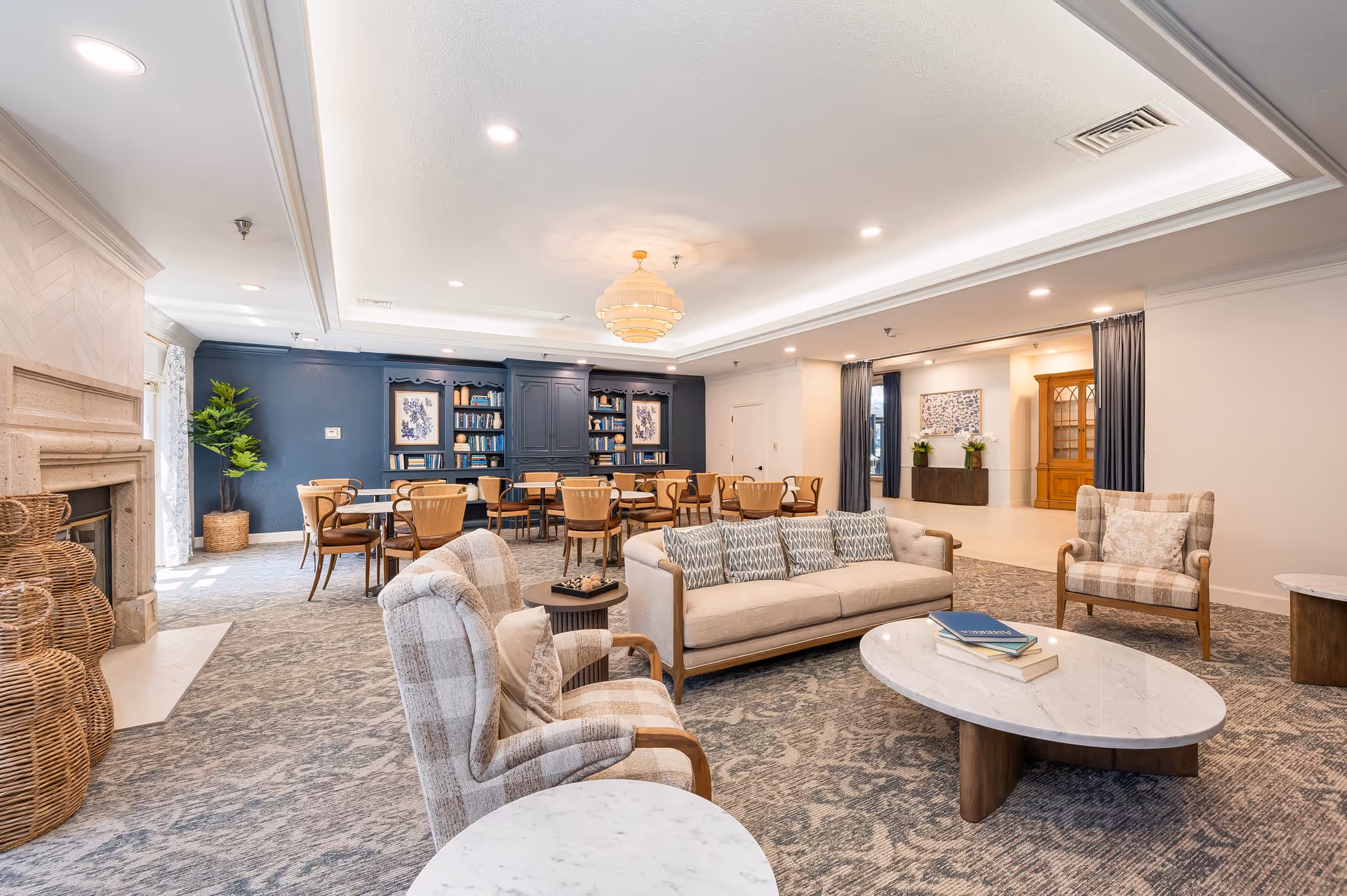 A spacious and well-lit common area featuring a beige sofa with patterned cushions, two plaid armchairs, and marble-topped coffee tables. In the background, there are several wooden chairs and tables arranged near a dark blue wall with built-in bookshelves and framed artwork. The room has a decorative ceiling light fixture, a fireplace on the left side, and light-colored walls and carpet.