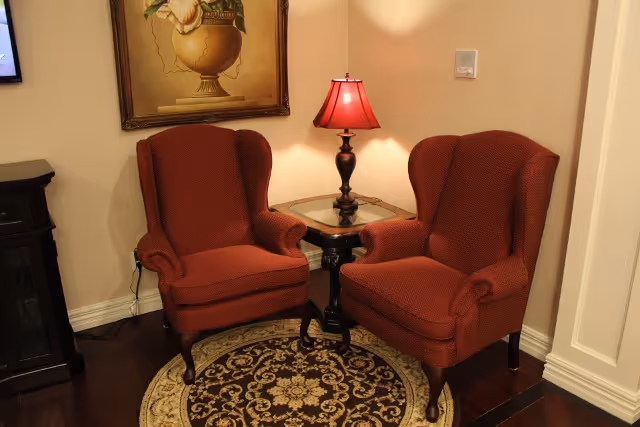 Two red upholstered armchairs positioned around a small wooden side table with a red lampshade, placed on a round patterned rug in a warmly lit corner of a room with beige walls and a framed painting above one chair.